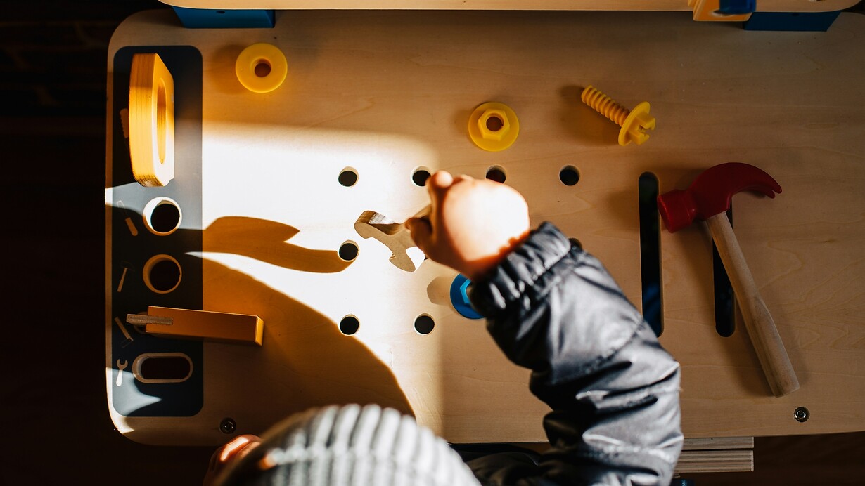 Kind an einer Spielwerkbank aus Holz, das mit einem Spielzeughammer und verschiedenen Schrauben und Muttern hantiert.